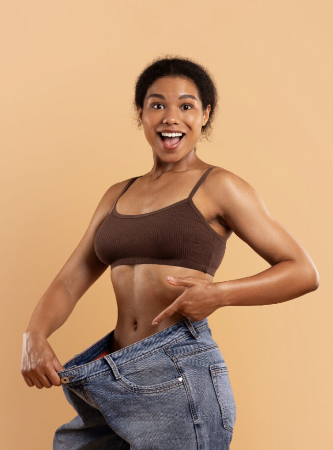 A fit African American woman smiling confidently. Used self hypnosis. She is showing her now over sized jeans, after successful fat loss hypnosis to remove belly fat.