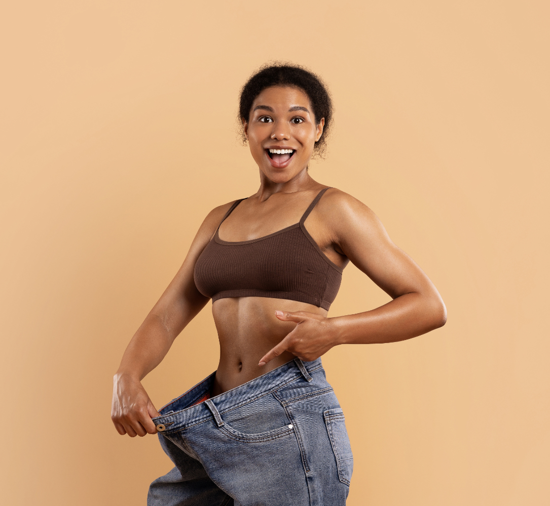 A fit African American woman smiling confidently. Used self hypnosis. She is showing her now over sized jeans, after successful fat loss hypnosis to remove belly fat.